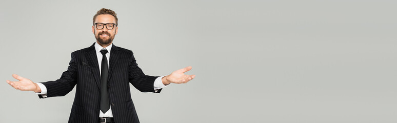 happy businessman in suit and glasses looking at camera and showing welcoming gesture isolated on...