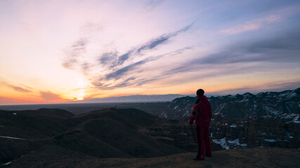 The guy in red is walking to the edge of the canyon. Red-orange light from the rising sun. Purple clouds. Orange canyon Charyn. Morning. There is snow in places. A trip through the canyon. Kazakhstan