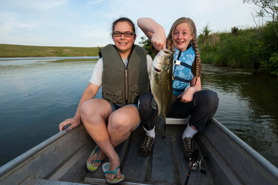 Two teenage girls fish from a boat on a farm pond, one holding their fresh catch of Largemouth bass (Micropterus salmoides); Valparaiso, Nebraska, United States of America