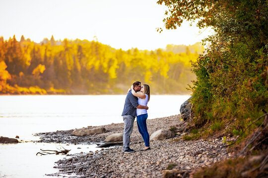 Married couple stand in an embrace and share a kiss beside a lake with autumn colours in the forest in the distance; Edmonton, Alberta, Canada