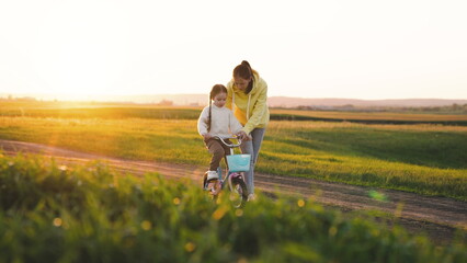 Naklejka na ściany i meble mother rides child bicycle sunset. happy family park. Mom teaches her daughter ride bike. concept happy family summer park. fun vacation. teamwork. mother runs green grass rolling child bicycle