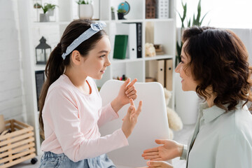 Side view of preteen girl gesturing and talking to smiling speech therapist in consulting room.