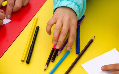 close-up of a child's hands drawing with colored pencils on a white sheet of paper on a red yellow table