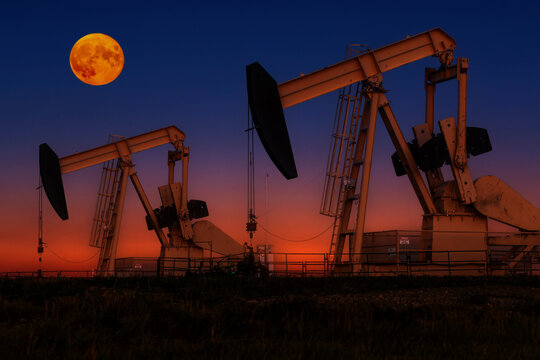 Glowing colourful pumpjacks at sunrise with glowing sky and full moon in the background, West of Airdrie; Alberta, Canada