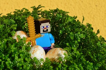 Naklejka premium LEGO Minecraft figure of Steve with golden sword standing in nest made of Selaginella plant, also called spike moss, with white golden stained easter eggs. Yellow wall in background.
