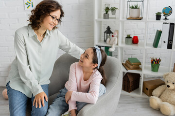 Cheerful psychologist in eyeglasses looking at happy preteen patient in consulting room.