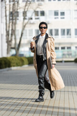 full length of african american businesswoman in black leather pants and trench coat holding laptop and paper cup while walking on blurred street.