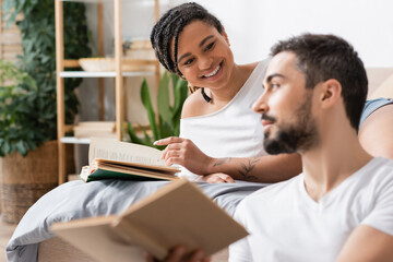 cheerful african american woman pointing at book near blurred boyfriend in bedroom at home.