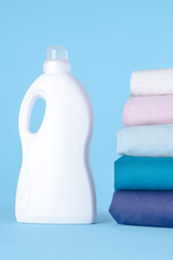 White plastic bottle for liquid laundry detergent, cleaning agent, bleach or fabric softener on blue background. Package mockup. A stack of colored laundry. Natural fabrics