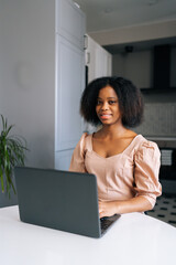 Vertical portrait of positive black female freelancer sitting at table in modern kitchen room working on laptop, smiling looking at camera. Happy African woman doing online job from home office.