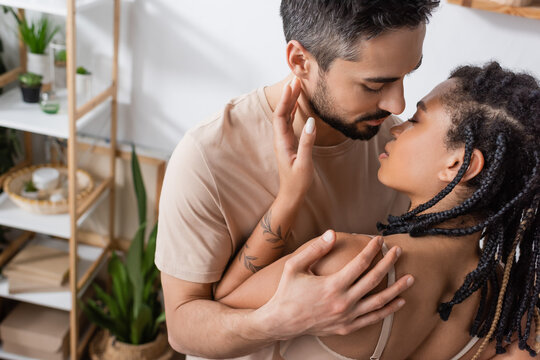 bearded brunette man in t-shirt hugging seductive african american woman with dreadlocks in bedroom at home.