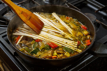 A wooden spatula mixes udon with stewed vegetables in spices in a frying pan.