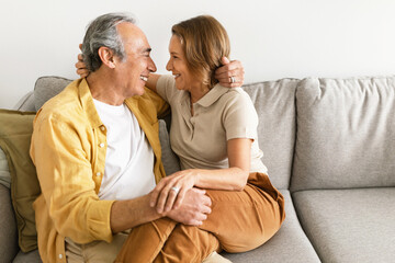Loving married senior spouses hugging, looking at each other and smiling, resting on sofa at home, free space