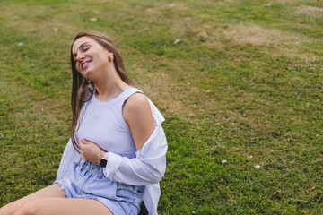 Nice looking woman is sitting on the grass in park, lookin at side and dreaming. She is relaxing, enjoying of the day. Girl look sensual and tenderness. Long hair female wear white shirt and top.