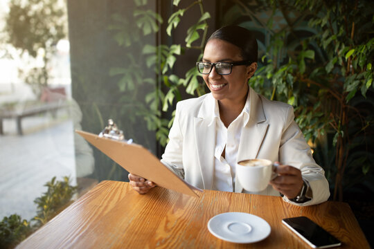 Glad millennial african american businesswoman in white suit and glasses drinking coffee and reading documents - Powered by Adobe