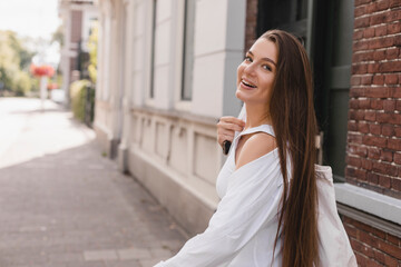 Attractive young brunette woman with long hair walk outdoor, cross road with shopping bag on shoulder. Girl turn around and laughing, good mood day. Female wear white shirt and blue jeans short.