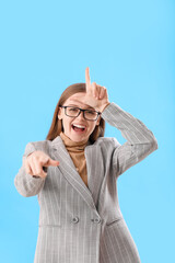 Young businesswoman showing loser gesture and pointing at viewer on blue background