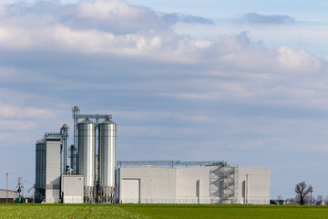 An animal feed production plant against the background of green agricultural fields. Grain silos next to the processing plant. Photo taken on a sunny day. © Fotoforce