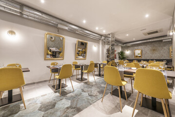 Dining room of a restaurant with assembled tables and chairs upholstered in yellow fabric and exposed air conditioning pipes