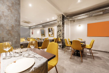 Dining room of a restaurant with tables, with cutlery, crockery, glassware and assembled chairs upholstered in yellow fabric and exposed air conditioning pipes