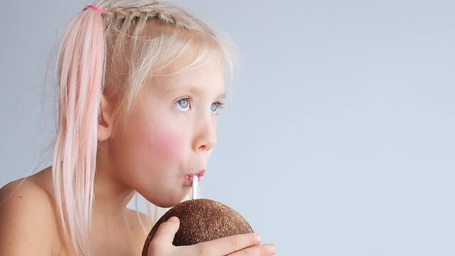 A little blonde girl drinks coconut water from coconut through a straw. Monochrome background. Close-up