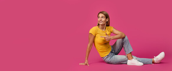 Positive middle aged european blonde lady sitting on floor pointing finger at empty space