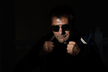 Mid aged person wearing sunglasses in a dark room holding his fists in a pose suggesting readiness to fight