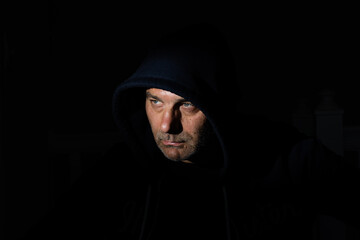 In a dimly lit room, a man in his fifties with stubbly, unshaven facial hair is wearing a hood that...