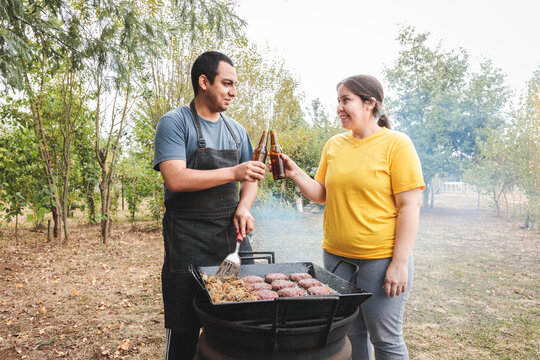 Latin young couple making a toast with beers while cooking homemade burgers on barbecue grill.