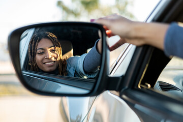 Dark-skinned young girl with braids sitting in a car looking in the rearview mirror while smiling....