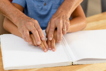 Cropped hands of caucasian grandfather assisting blind grandson in reading braille book at home