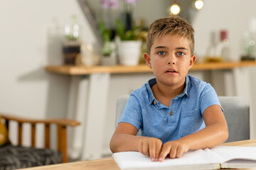 Portrait of caucasian blind boy reading braille book on table while sitting at home, copy space