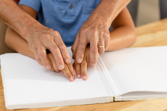 Cropped hands of caucasian grandfather assisting blind grandson in reading braille book at home - Powered by Adobe