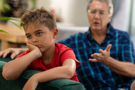 Caucasian sad boy with hand on chin looking away while sitting with grandfather on sofa