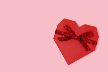 Red gift box in a heart shape with tied glittering ribbon. Minimal festive concept on a pink background.