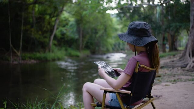 A woman using a digital tablet while sitting on a camping chair near the river
