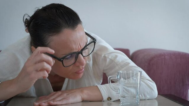 Glass half empty half full on table. A pensive woman in glasses sits on the sofa and think about half full or half empty glass on the table.