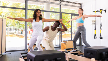 Fototapeta na wymiar Attentive African American male instructor helping two positive women performing set of pilates exercises on reformer during group classes in modern health club