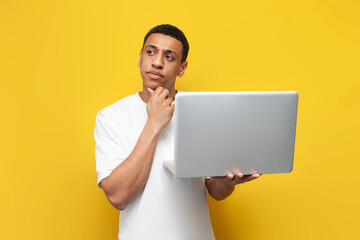 young pensive african american guy in white t-shirt uses laptop and dreams on yellow isolated...