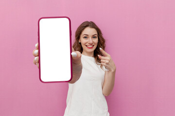 young cute woman in white t-shirt shows blank smartphone screen on pink isolated background