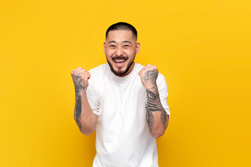 unshaven Asian man in white t-shirt rejoices in victory on yellow isolated background, adult guy...