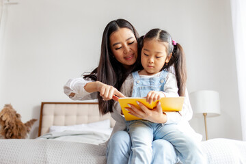 asian family sitting on bed and reading book, korean woman mother teaching little daughter to read book