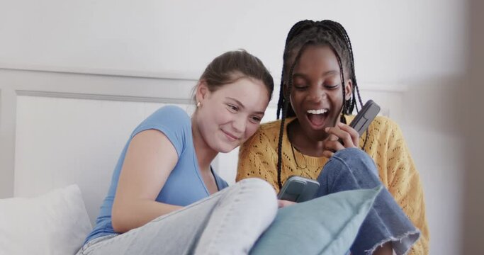 Happy diverse teenage female friends lying on bed using smartphones and laughing