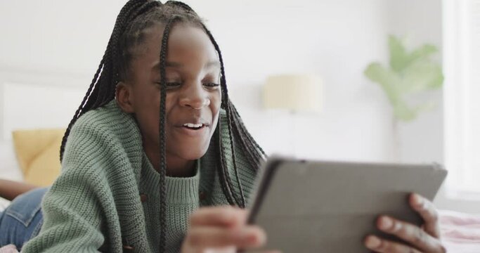 Happy african american teenage girl lying on bed using tablet and laughing