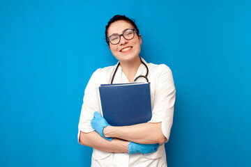 female doctor student in a medical gown holds documents and papers on a blue isolated background, a girl intern nurse