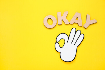 Fototapeta na wymiar Word Okay made of wooden letters and paper cutout (OK hand gesture) on yellow background, flat lay. Space for text
