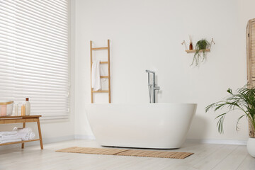 Stylish bathroom interior with ceramic tub, care products and terry towels on table