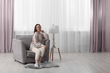 Fototapeta premium Woman sitting on armchair near lamp and window with stylish curtains at home. Space for text