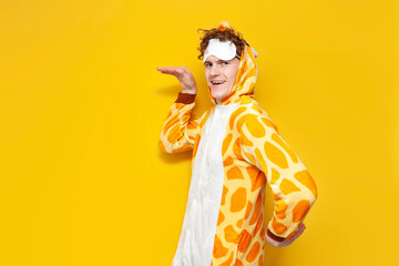 young joyful guy in funny baby giraffe pajamas and sleep mask is dancing on yellow background