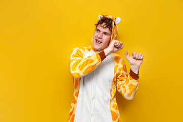 young joyful guy in funny children's giraffe pajamas dances on yellow background, man in animal...
